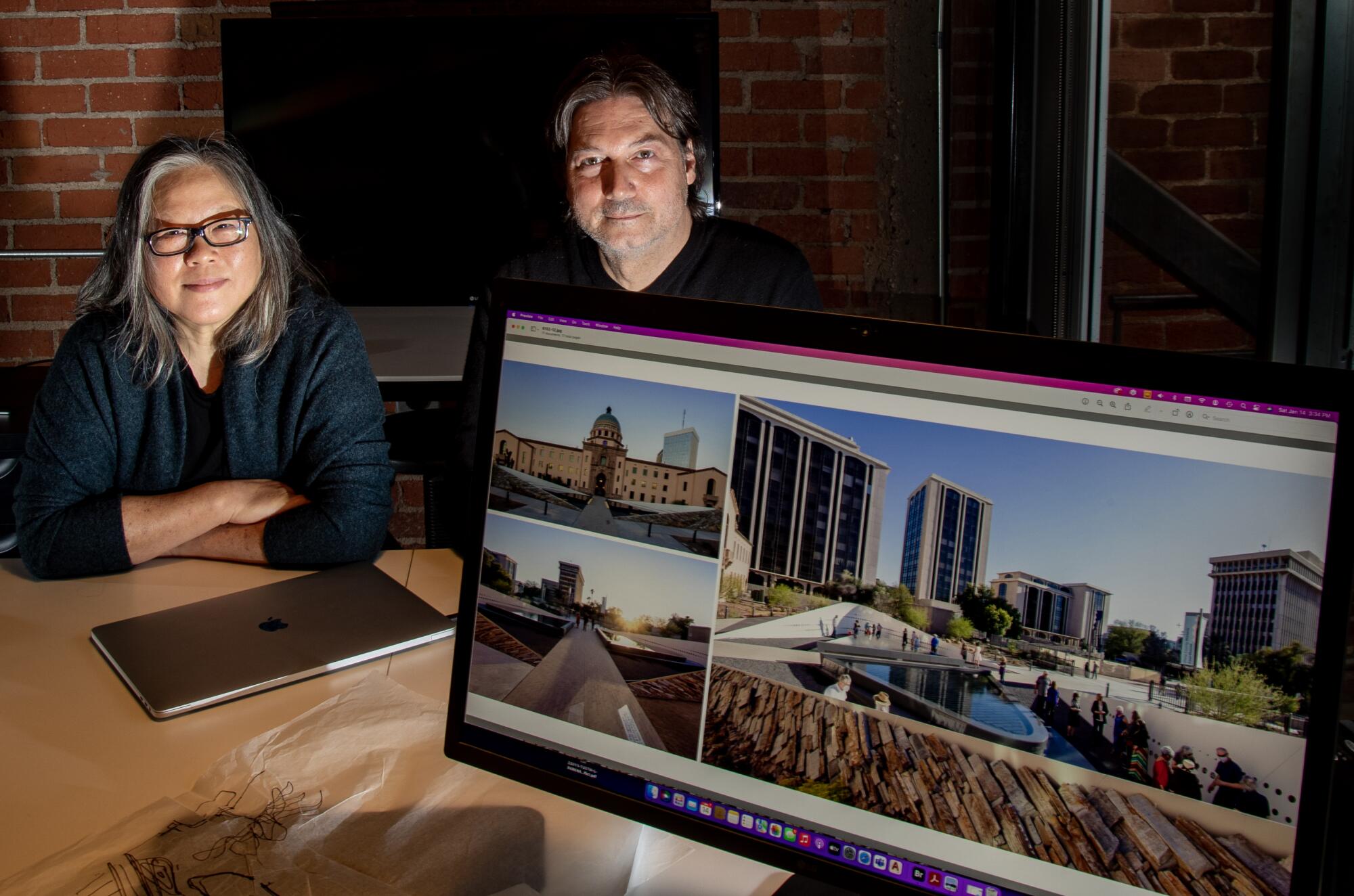 A woman and man sit at a wood table in moody light, behind a monitor bearing images of the Jan. 8 memorial.