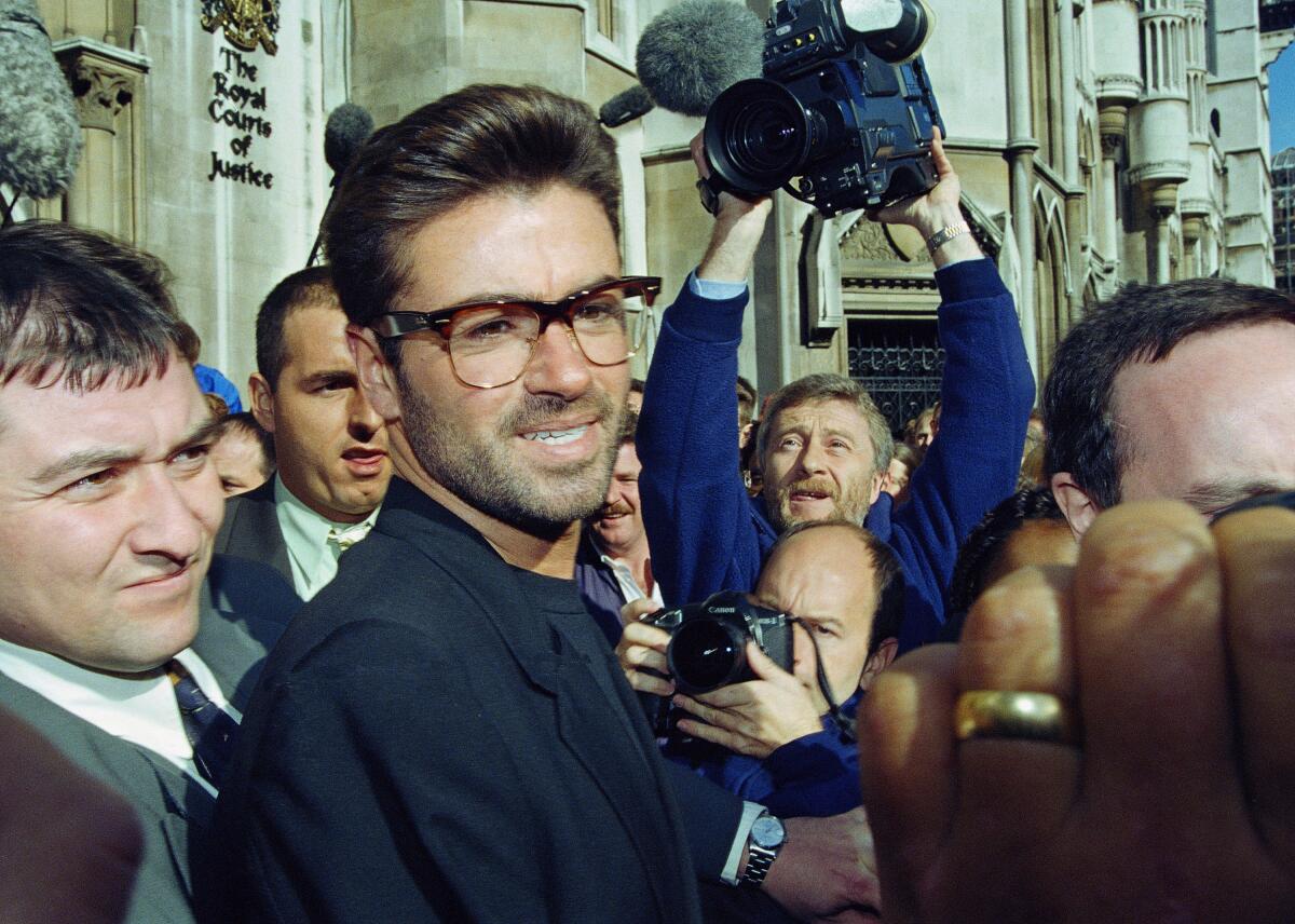A man with glasses stands amid a crowd of photographers and onlookers