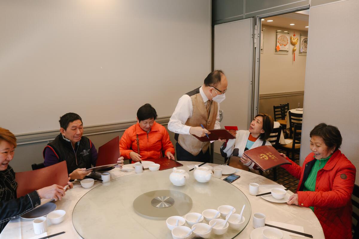 At center a waiter talks to a customer while five people are seated around the round table with white bowls and tea pots.