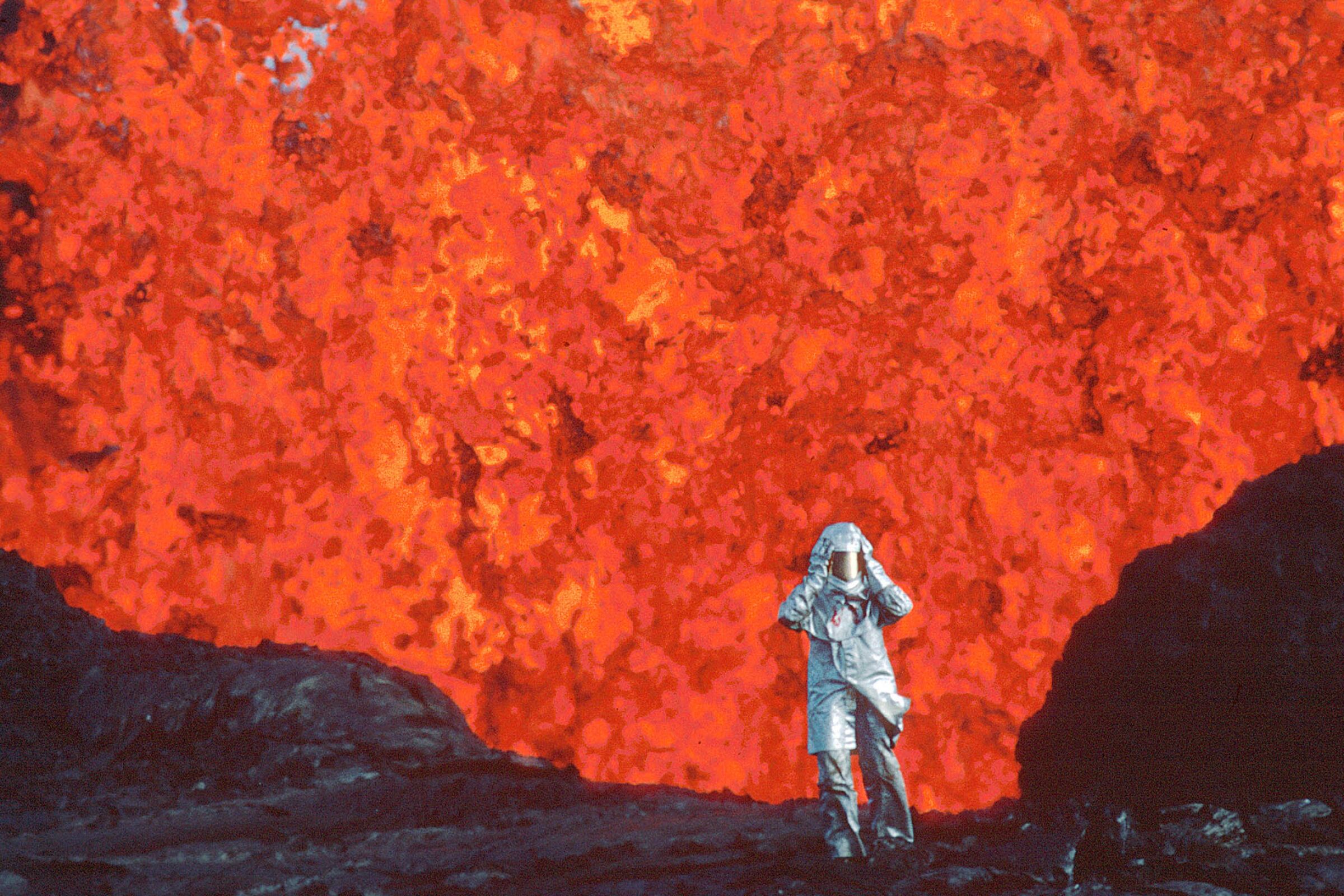 A figure in a silver fireproof suit and helmet stands on the edge of a volcano with lava raging behind.