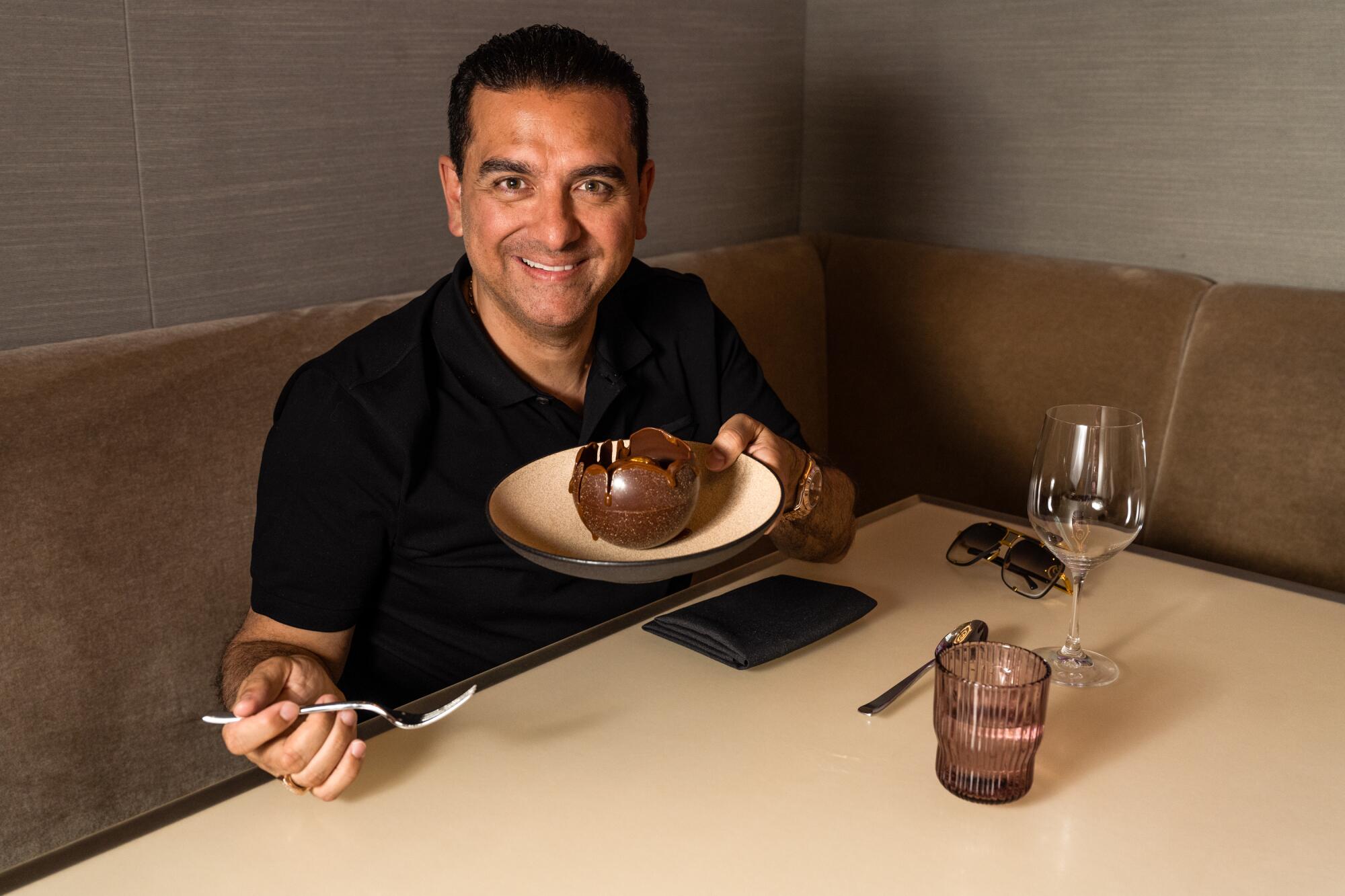 Buddy Valastro, seated at a restaurant table, holds up a plate with a chocolate-covered dessert