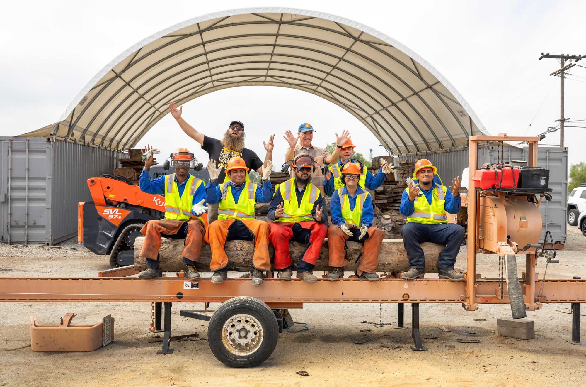 A group of people in orange safety helmets and yellow reflective vests sit on a log in a trailer and wave.