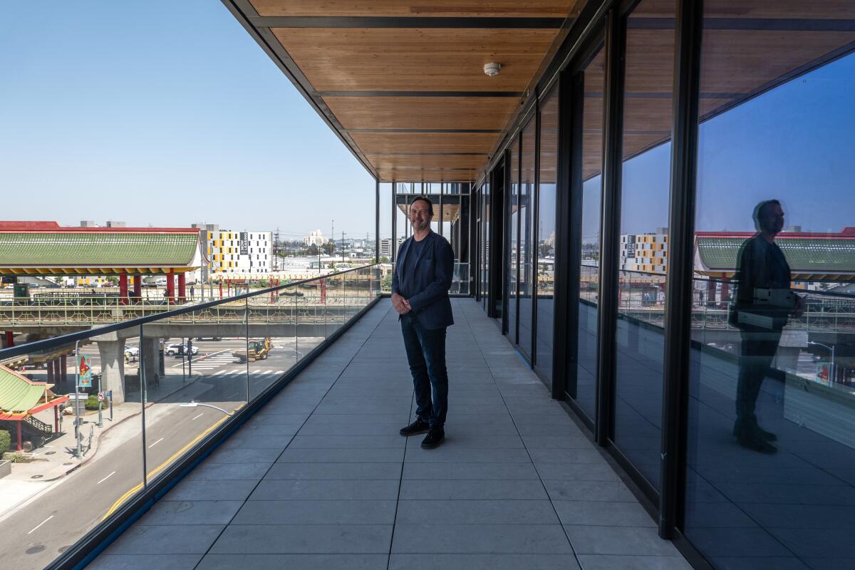 A man in a black suit stands on a deck of a building under construction, an elevated Metro stop visible in the distance.
