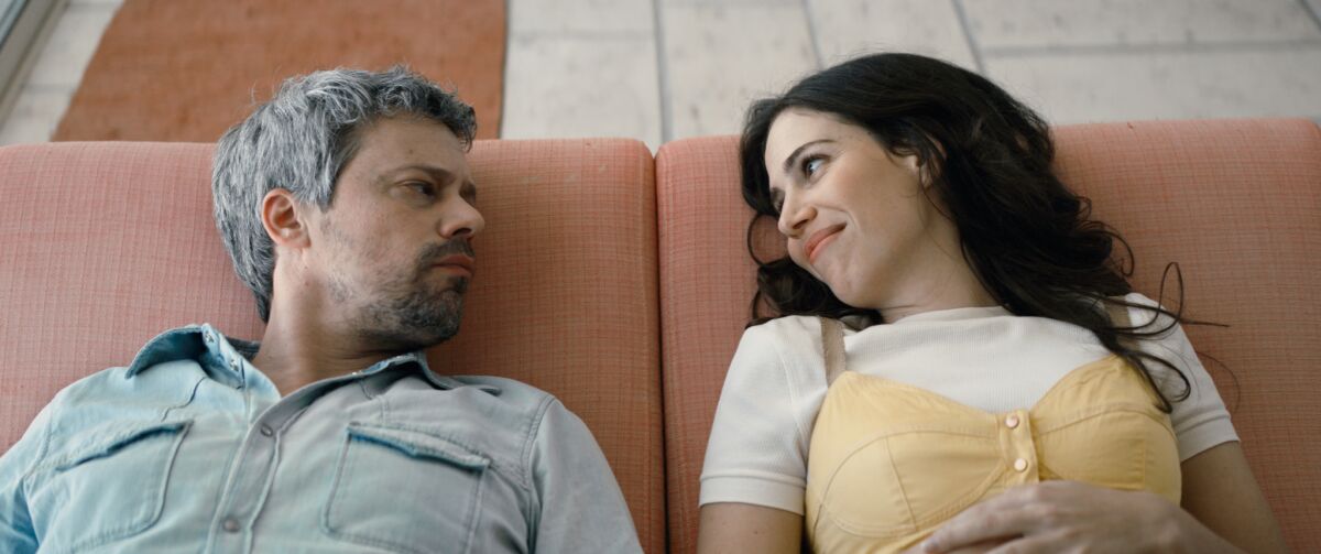 A man and woman look at each other as they recline side by side on a couch.