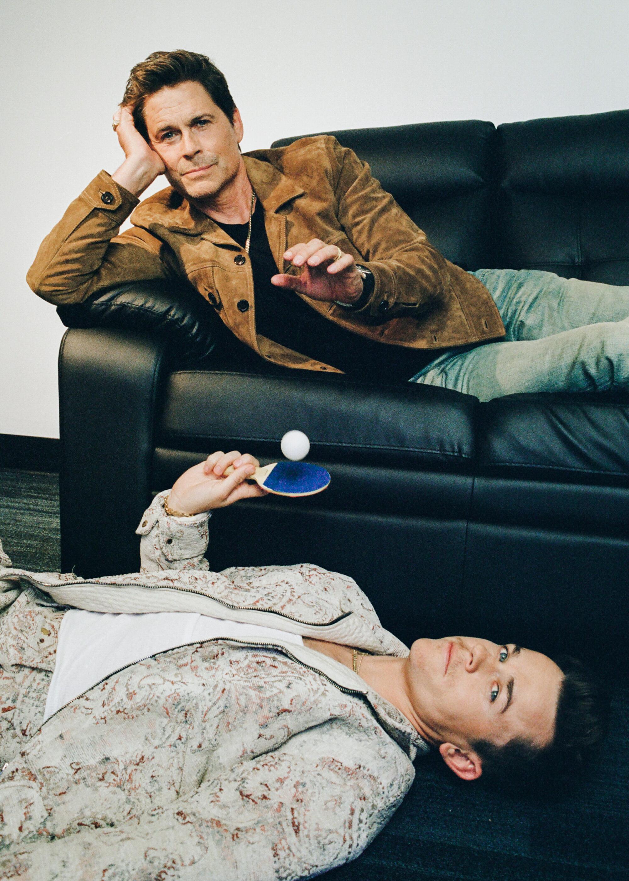 Rob Lowe lies on a couch and drops a ping pong ball on a paddle being held up by his son John Owen Lowe, lying on the floor.