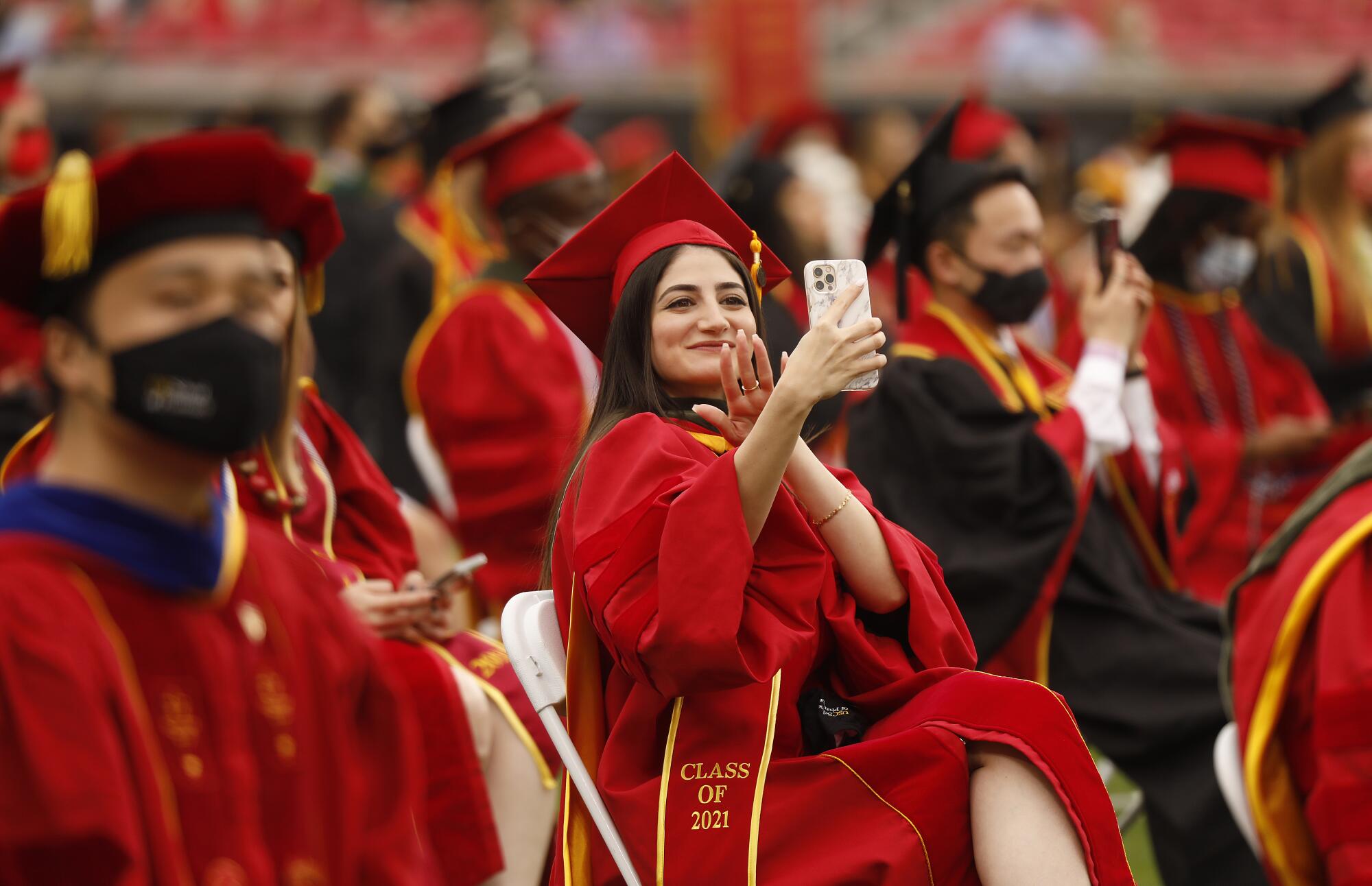 A woman in a red graduation gown smiles and waves at her phone while on a video call