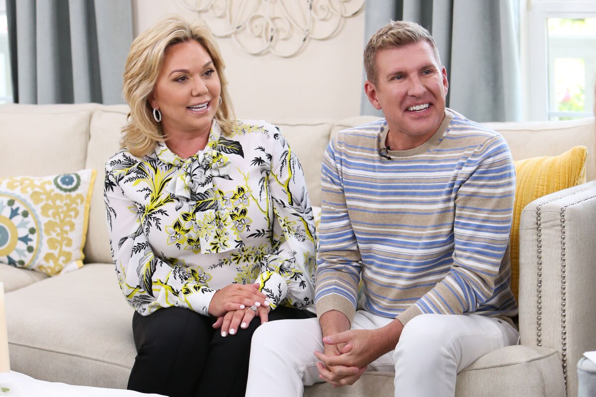 Woman dressed in floral blouse and man in blue and white long sleeve sit on gray couch in TV studio, smiling.
