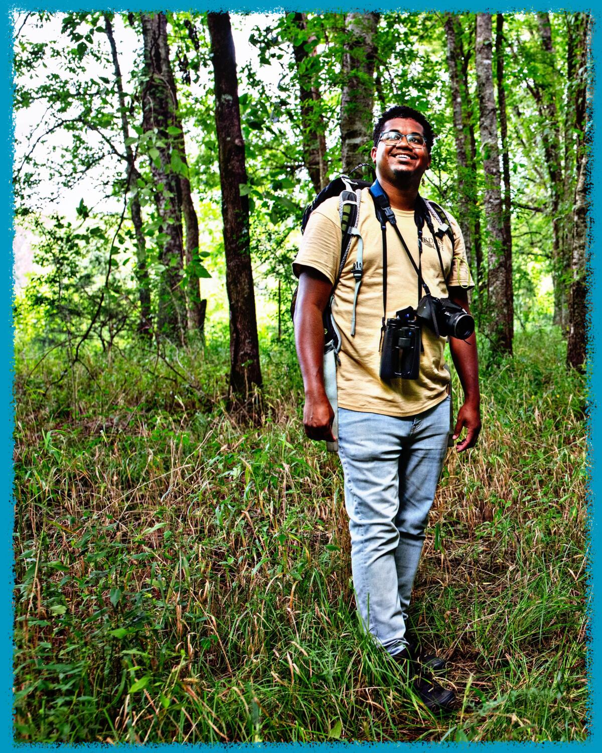 A man wearing a backpack, with a camera and binoculars slung around his neck, stands amid trees.