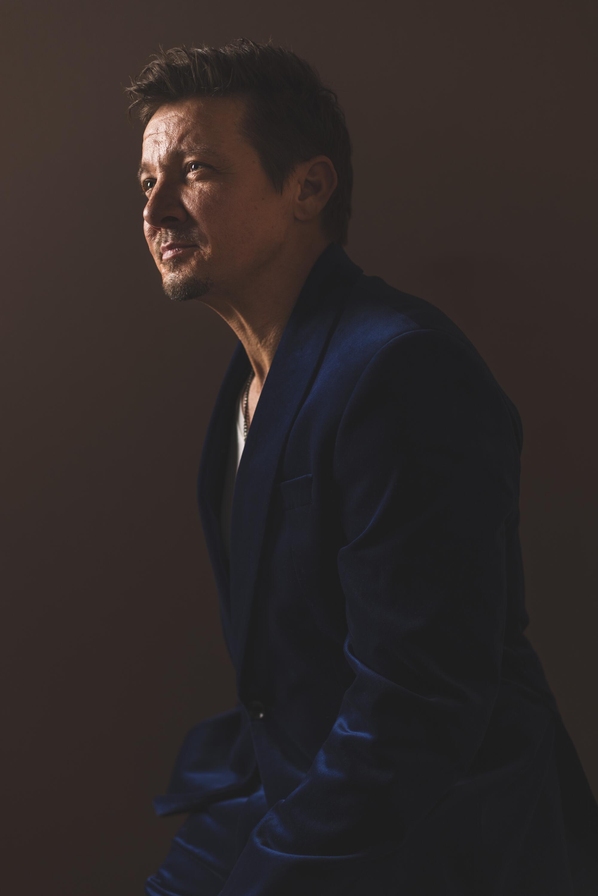 A smiling Jeremy Renner in a dark suit, leaning forward toward a light from above.