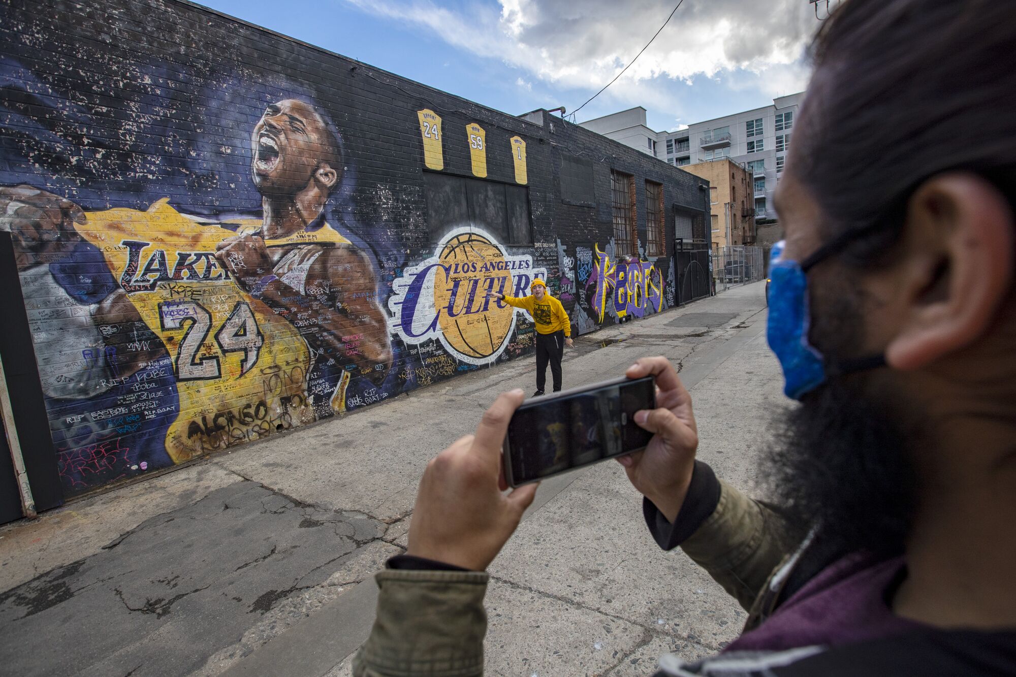 A man holds up his cellphone to take a picture of another man in front of a mural of Kobe Bryant in a Lakers uniform.