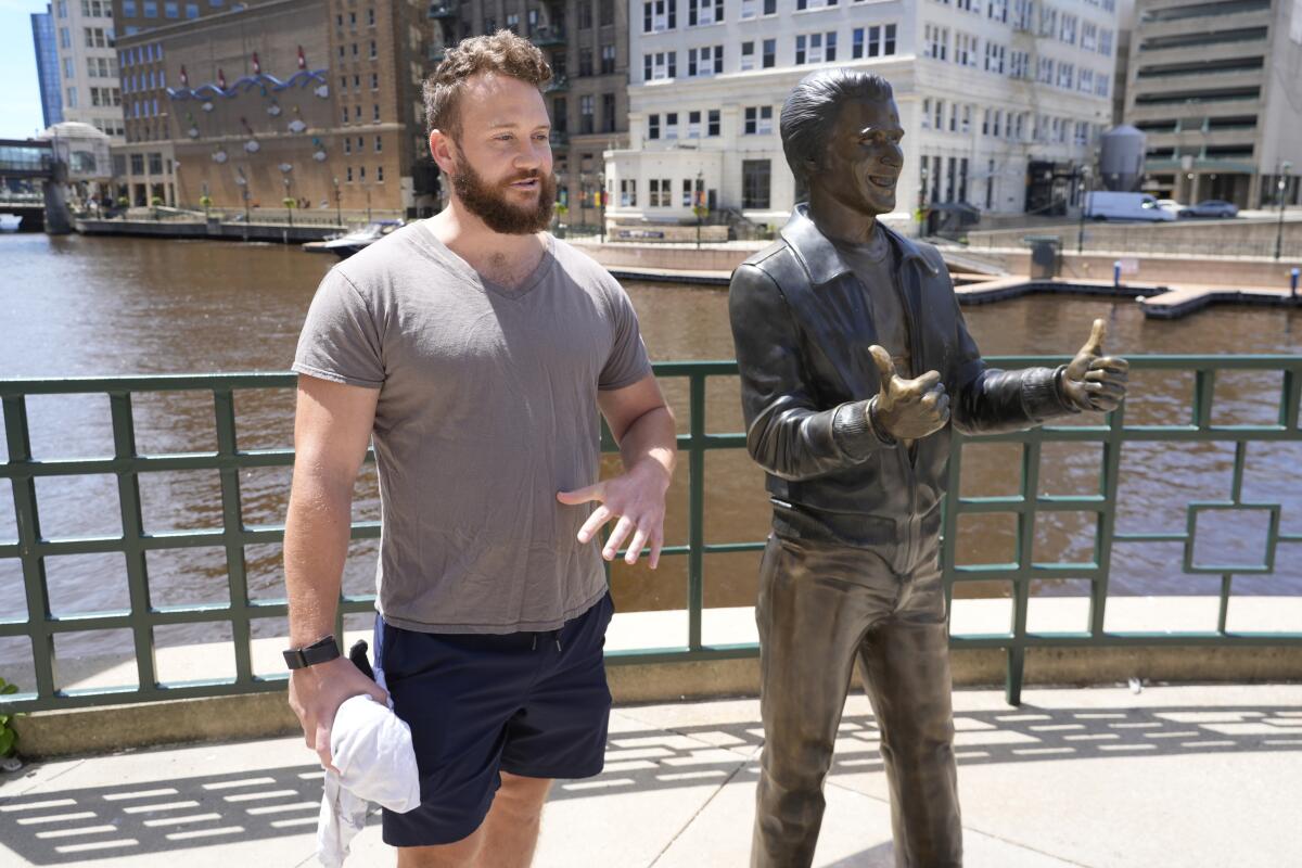 A man stands near a river behind a bronze statue of the character Fonzie. 