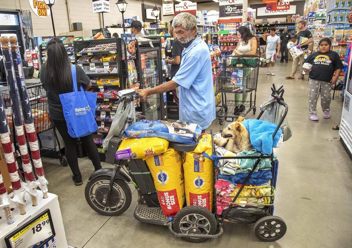 A man waits in a grocery store checkout line next to his scooter and cart that's filled with pet food and a dog. 