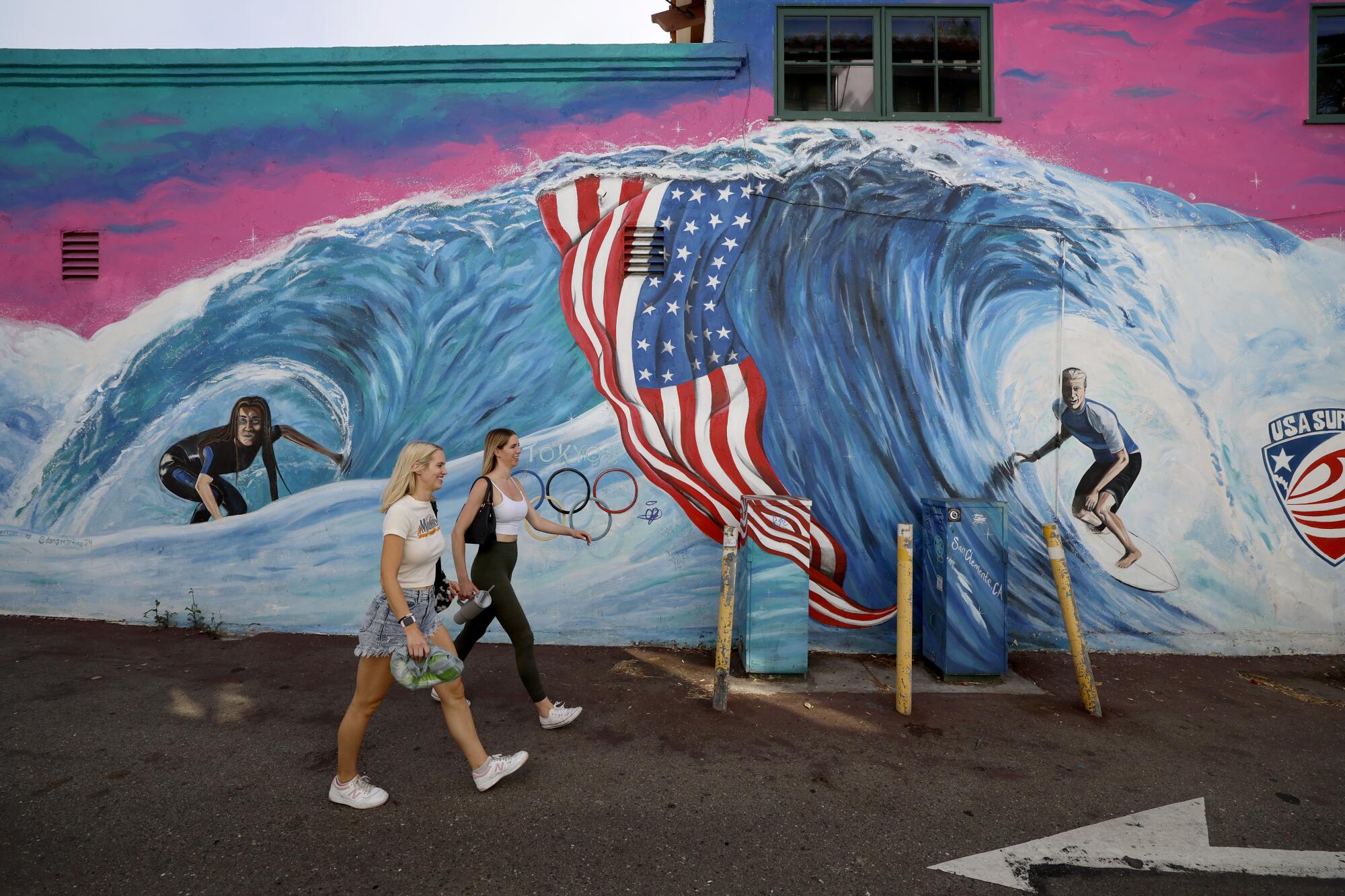 Young women walk past a mural depicting two Olympic surfers from San Clemente.