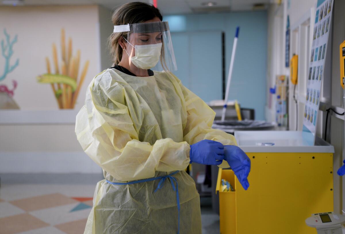 A hospital nurse wearing personal protective equipment, including a mask and face shield, puts on gloves