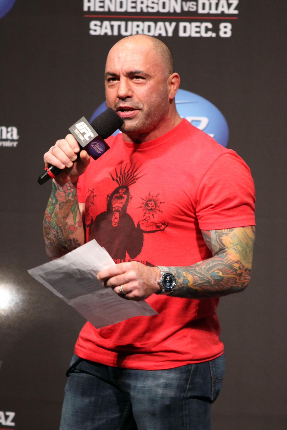 A bald man with a red T-shirt and arm tattoos speaks into a microphone