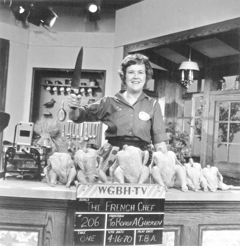 A woman stands behind a kitchen counter with six whole raw chickens; she holds up a large knife and smiles.