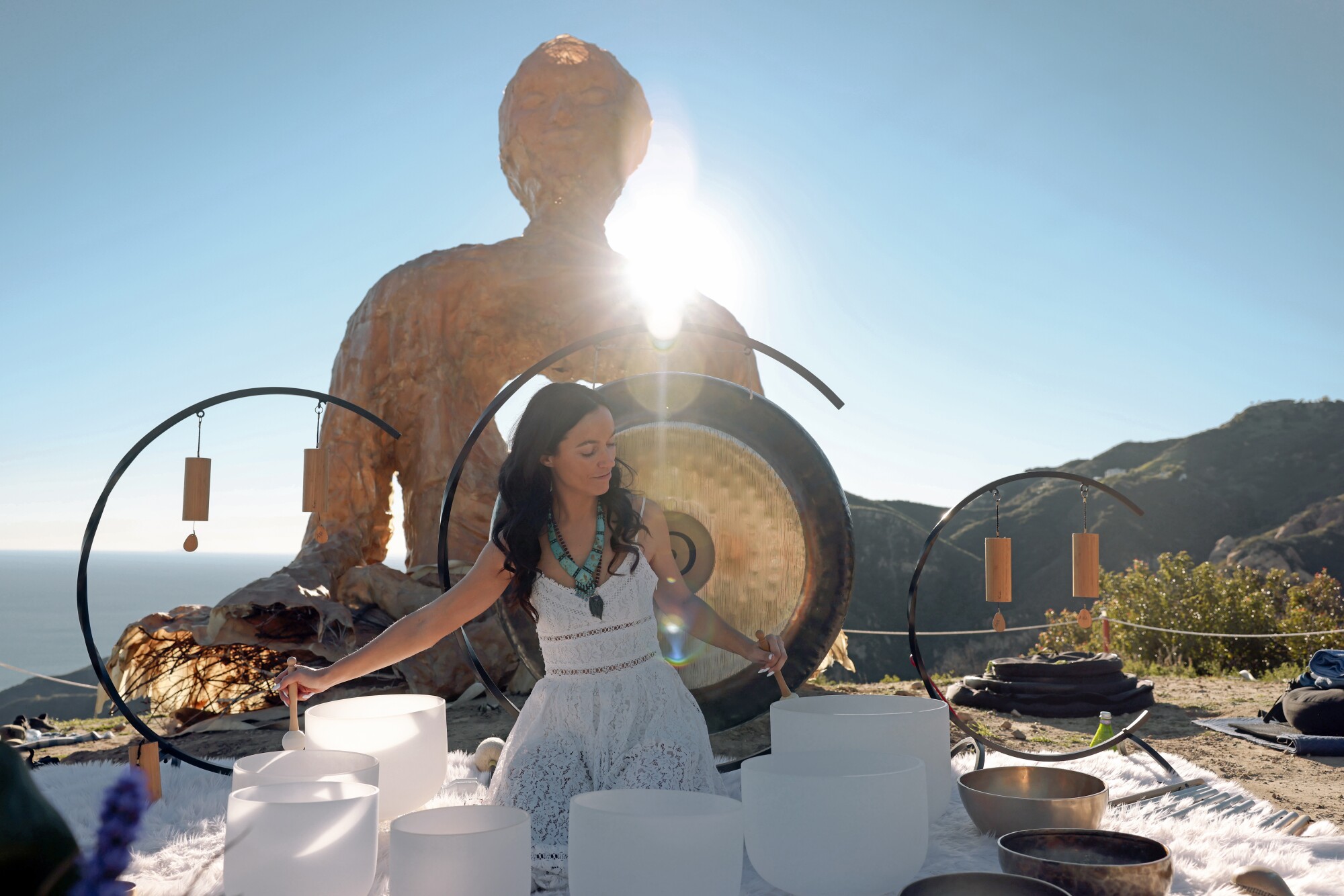 A woman in a white dress sits amid tuning bowls and gongs with the ocean in background.