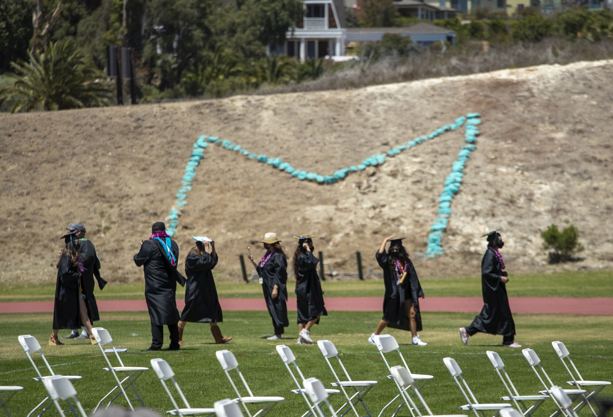Students walk in caps and gowns alongside rows of folding chairs. In the background, rocks form an "M" on a hillside.