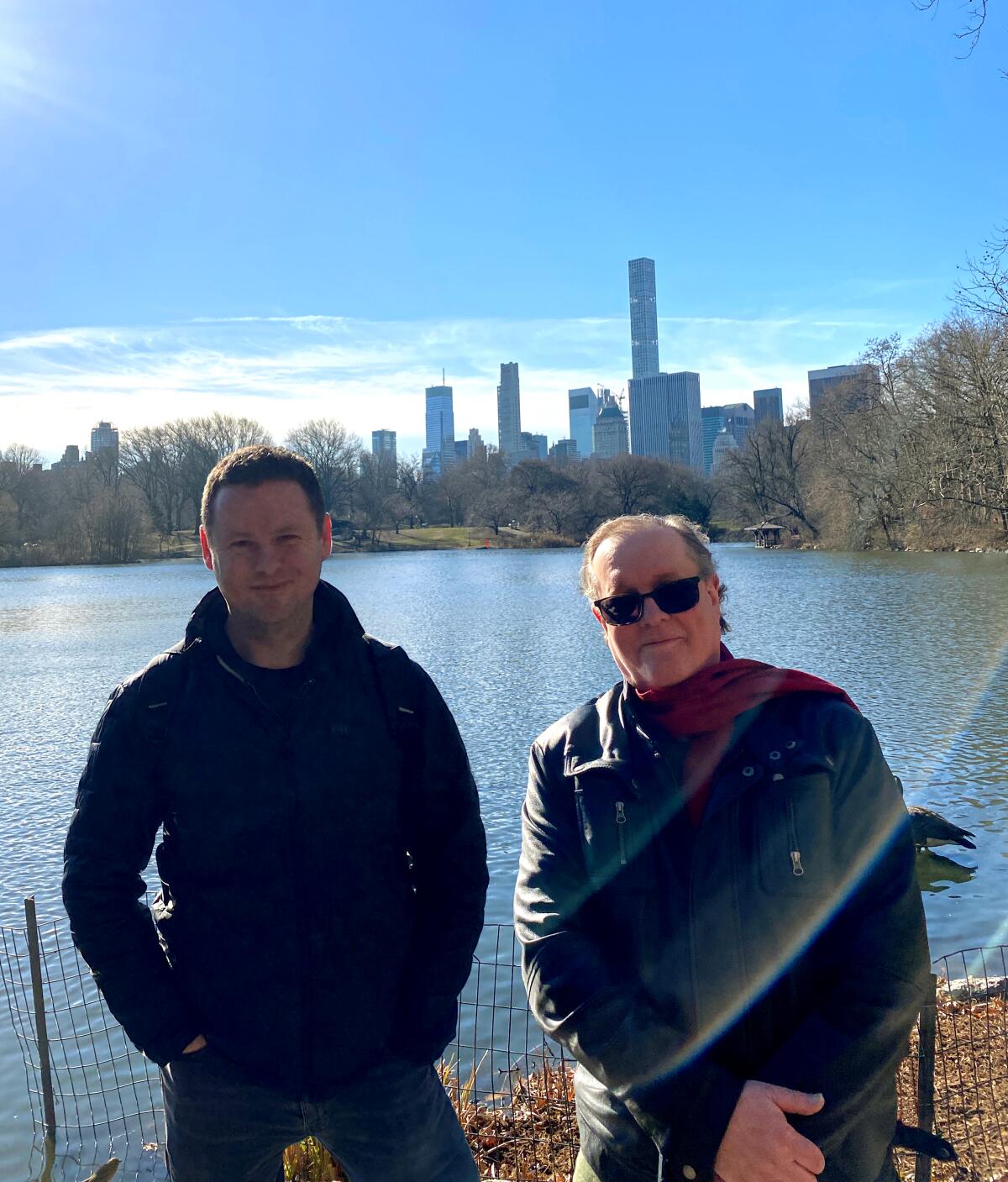 Two men pose in front of a lake and the New York skyline.