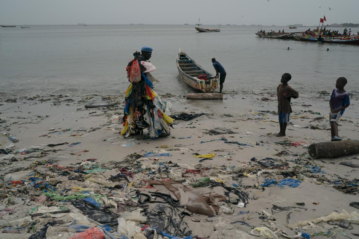 Kids look at environmental activist Modou Fall, who many simply call "Plastic Man", while he walks on the Yarakh Beach littered by trash and plastics in Dakar, Senegal, Tuesday, Nov. 8, 2022. As he walks, plastics dangle from his arms and legs, rustling in the wind while strands drag on the ground. On his chest, poking out from the plastics, is a sign in French that says, "No to plastic bags." (AP Photo/Leo Correa)