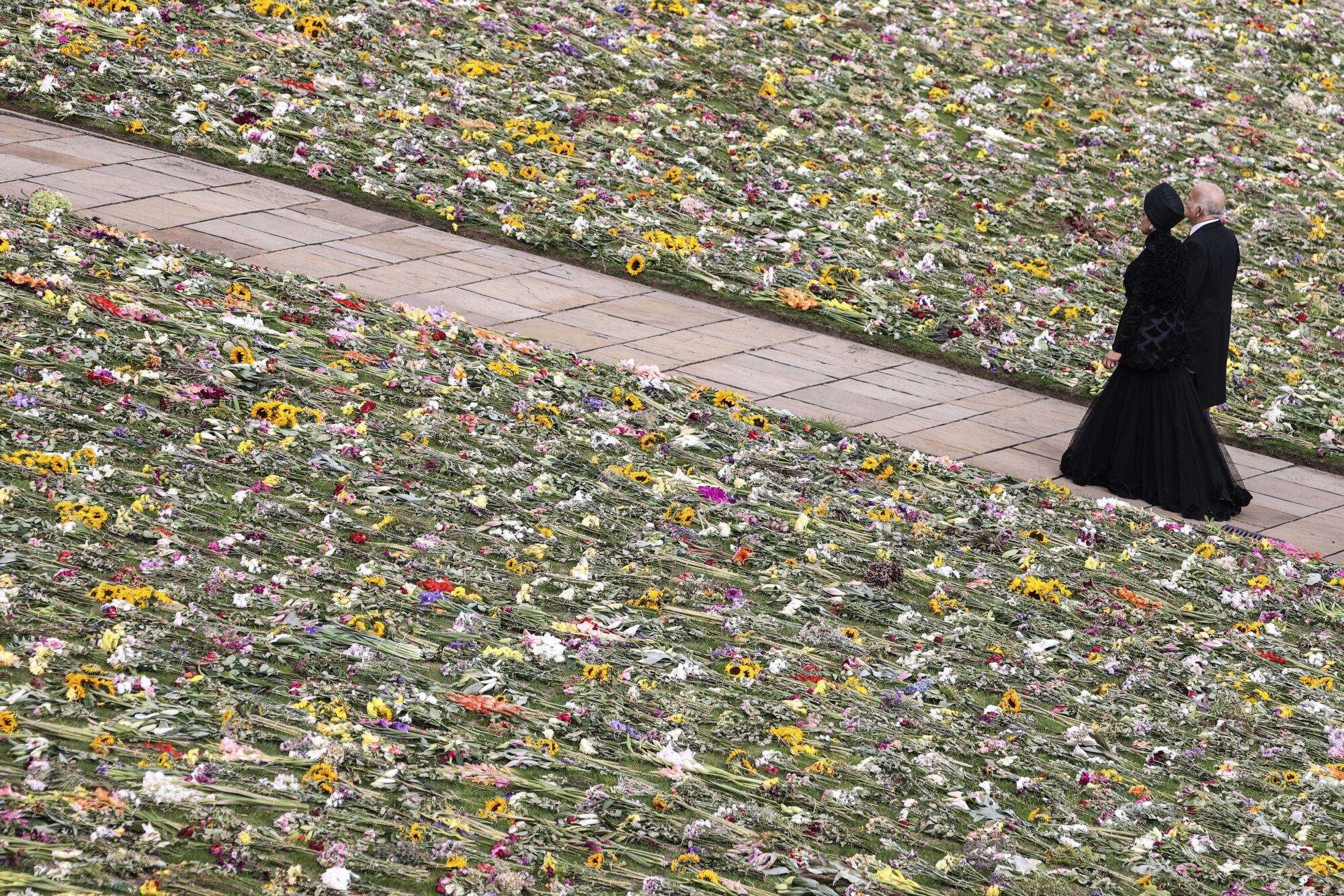 A man and woman dressed in black walk on a sidewalk past a lawn covered in flowers.