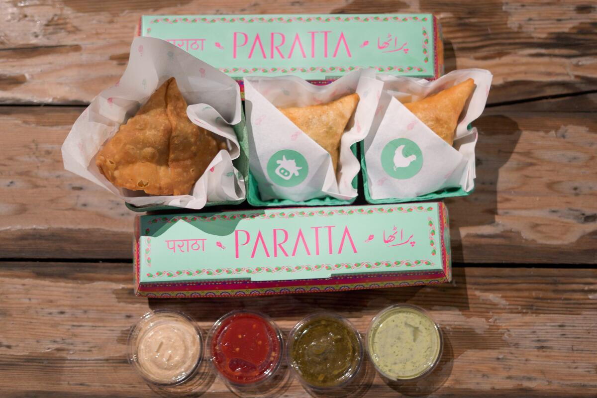 Three paper-wrapped fried samosas, with small plastic containers of sauces, in a colorful box that says "Paratta"