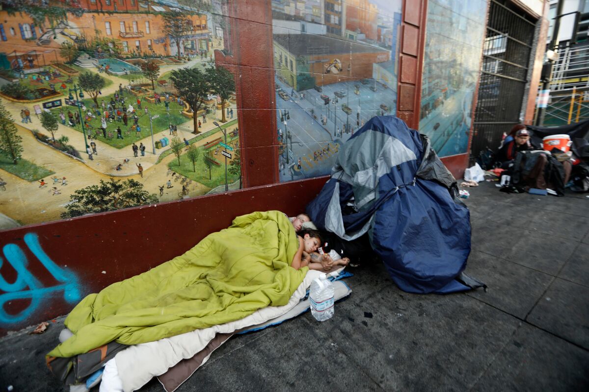 A man and a woman lie under blankets next to a tent on the sidewalk