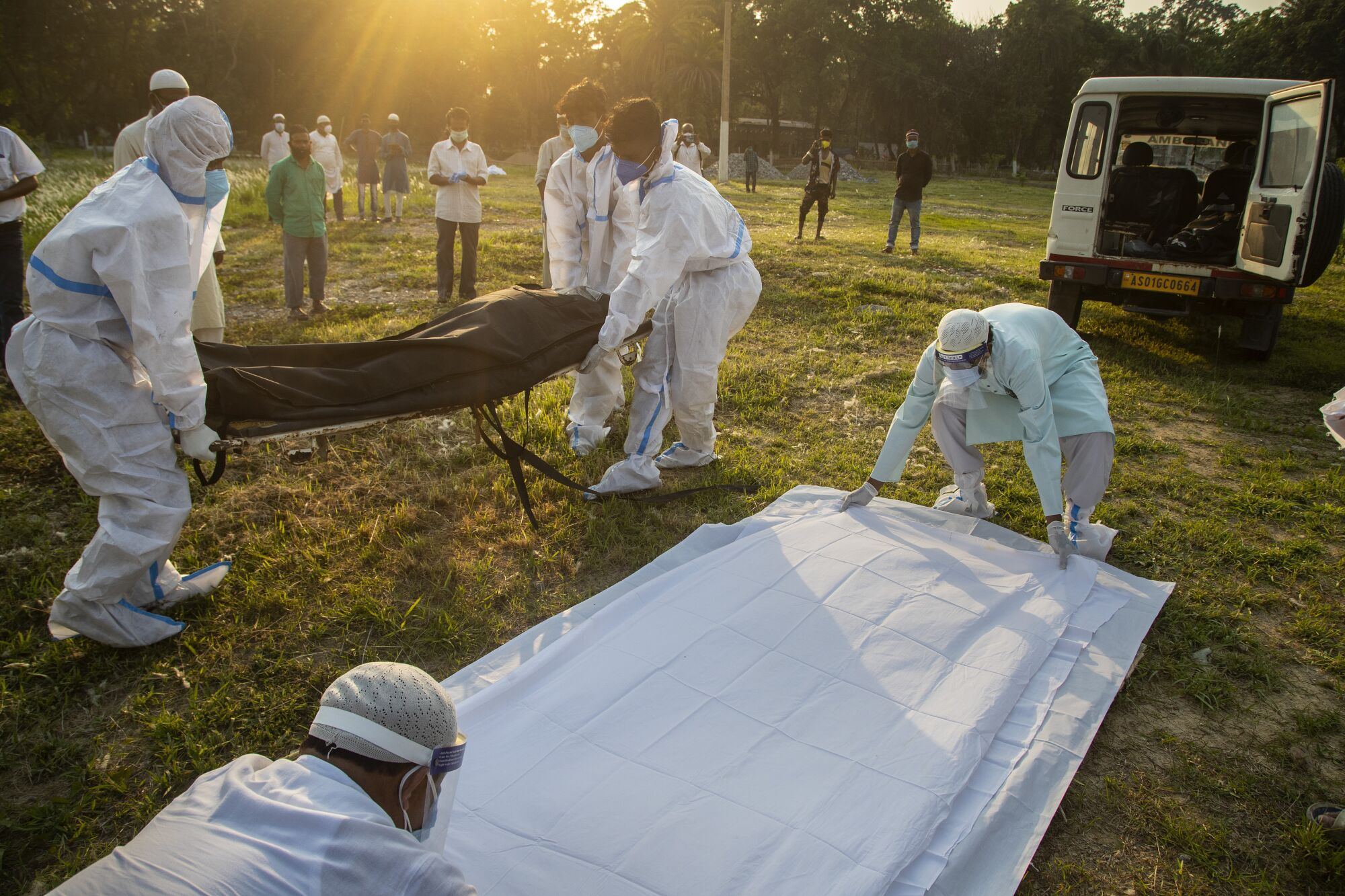 Workers in white protective suits, left, carry a black body bag. Two more workers lay out white sheets on the grass nearby.
