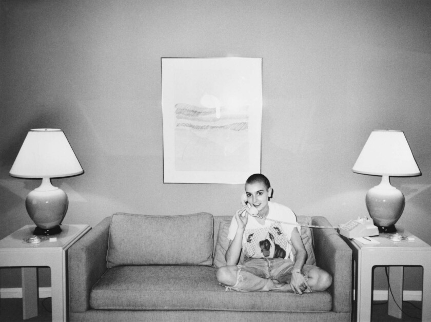 A photo of a young Sinéad O'Connor sitting on a couch between contemporary end tables and lamps.
