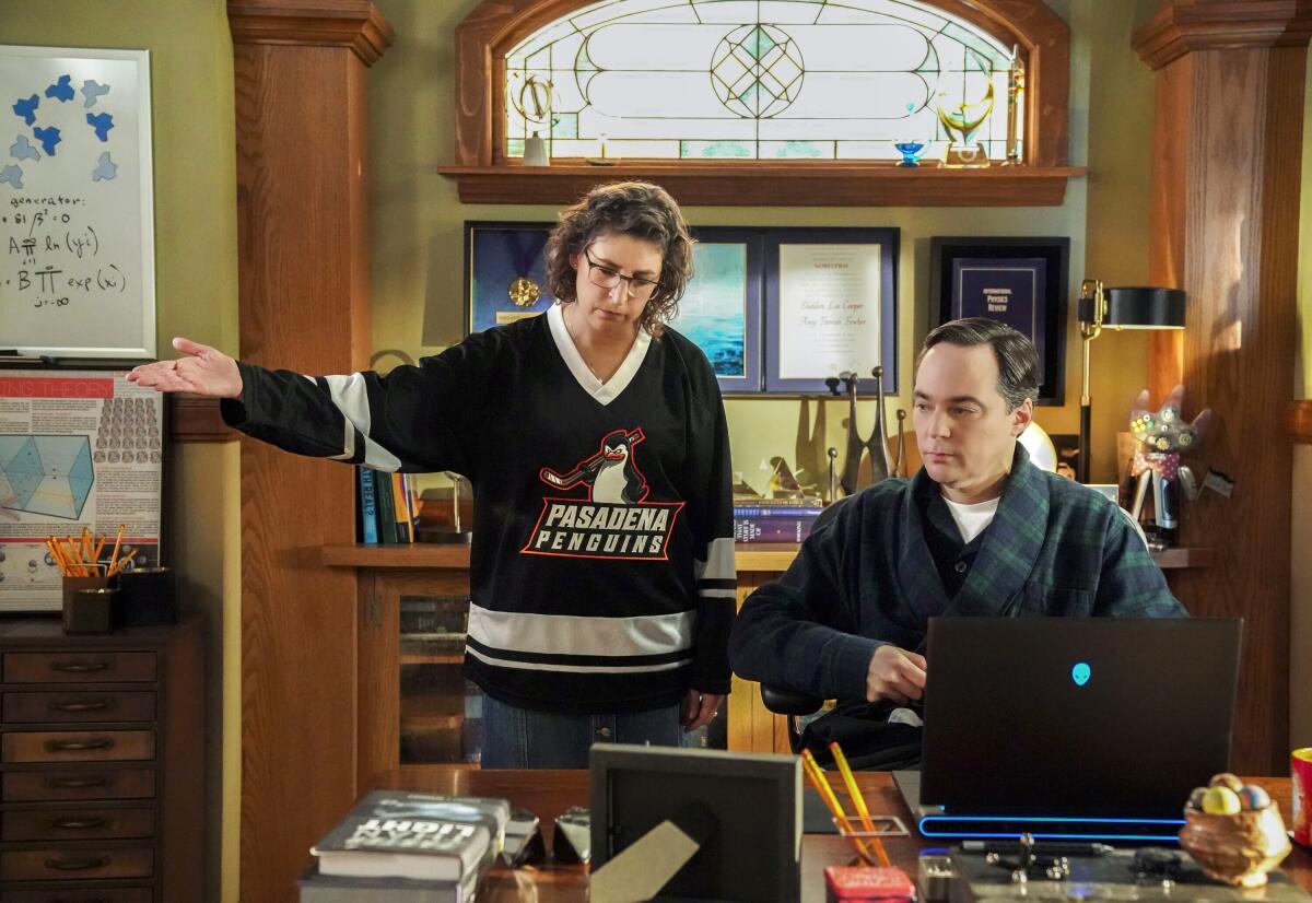 A woman in a hockey jersey stands with her arm out while looking at a man in a robe in front of a laptop.