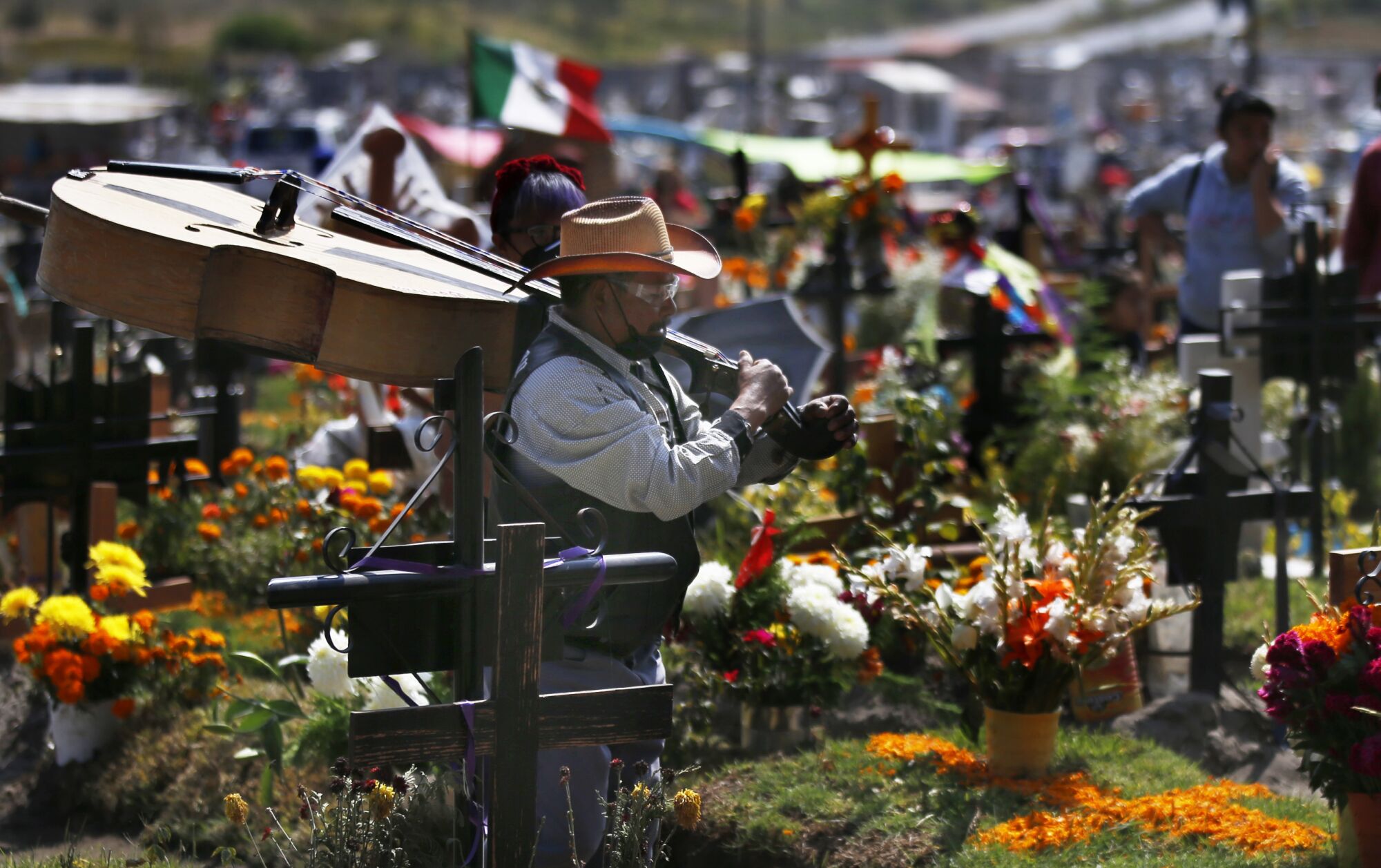 A musician carries a large upright bass over his shoulder while walking between flower-covered gravesites