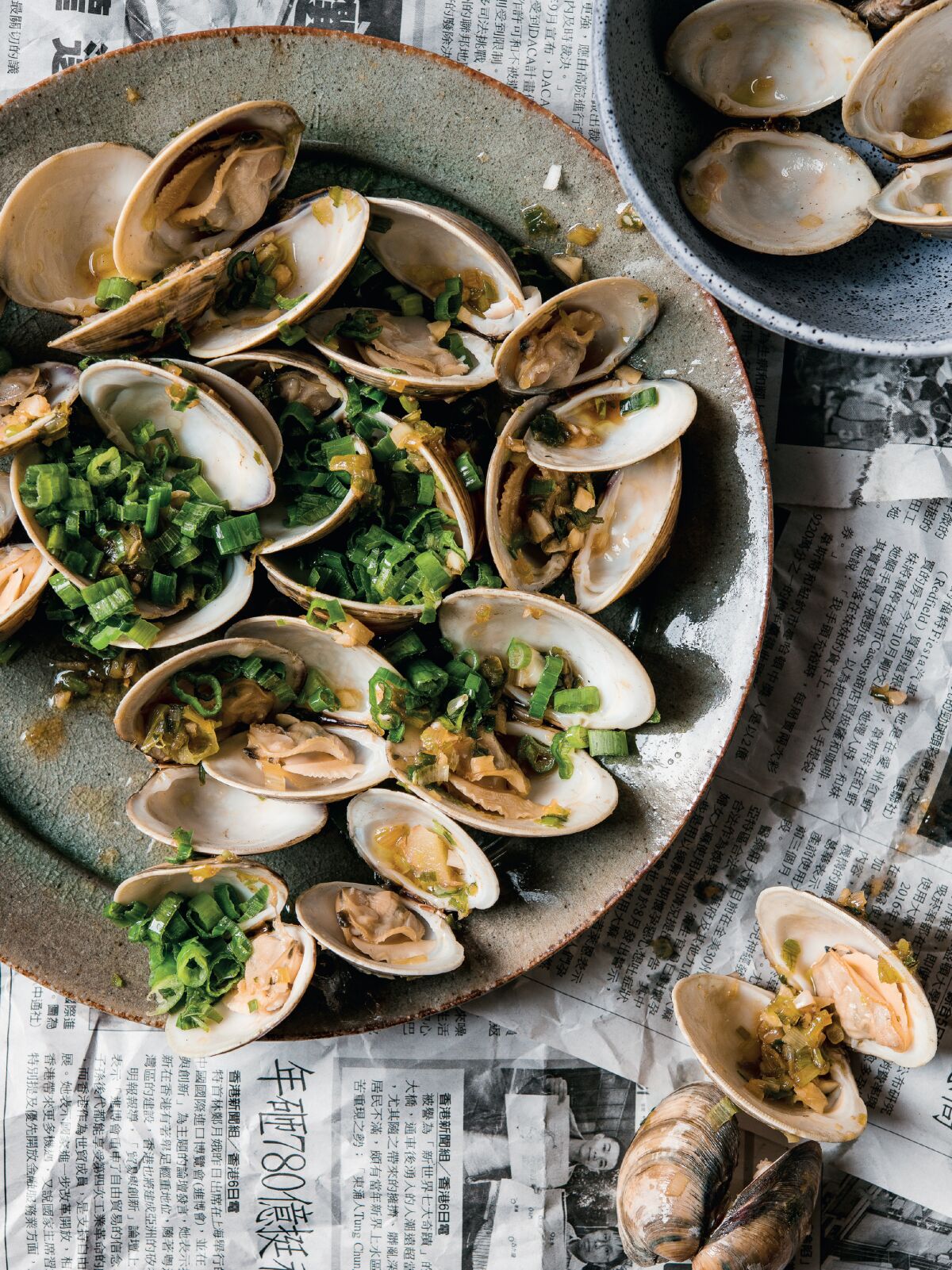 A plate filled clams and chopped scallions sits atop Chinese-language newspapers.