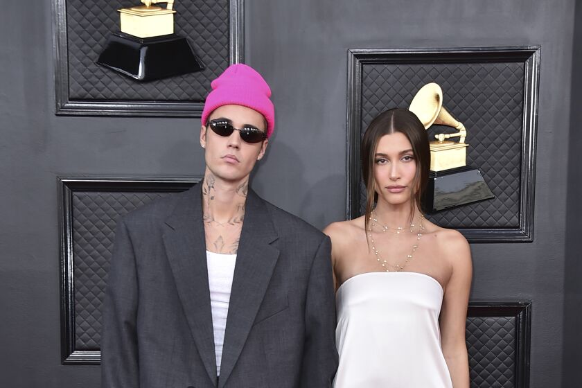 A man in an oversized suit and a pink beanie stands next to a woman in a white gown on a red carpet