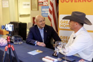 A man in a suit and a man in a cowboy hat sit next to each other at a table.