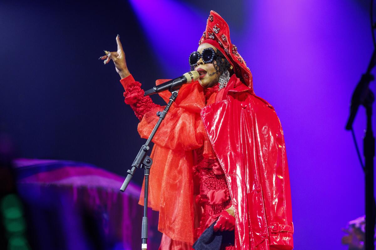 Lauryn Hill clad in a red latex coat with a hood and large sunglasses, sings and points up with right hand.