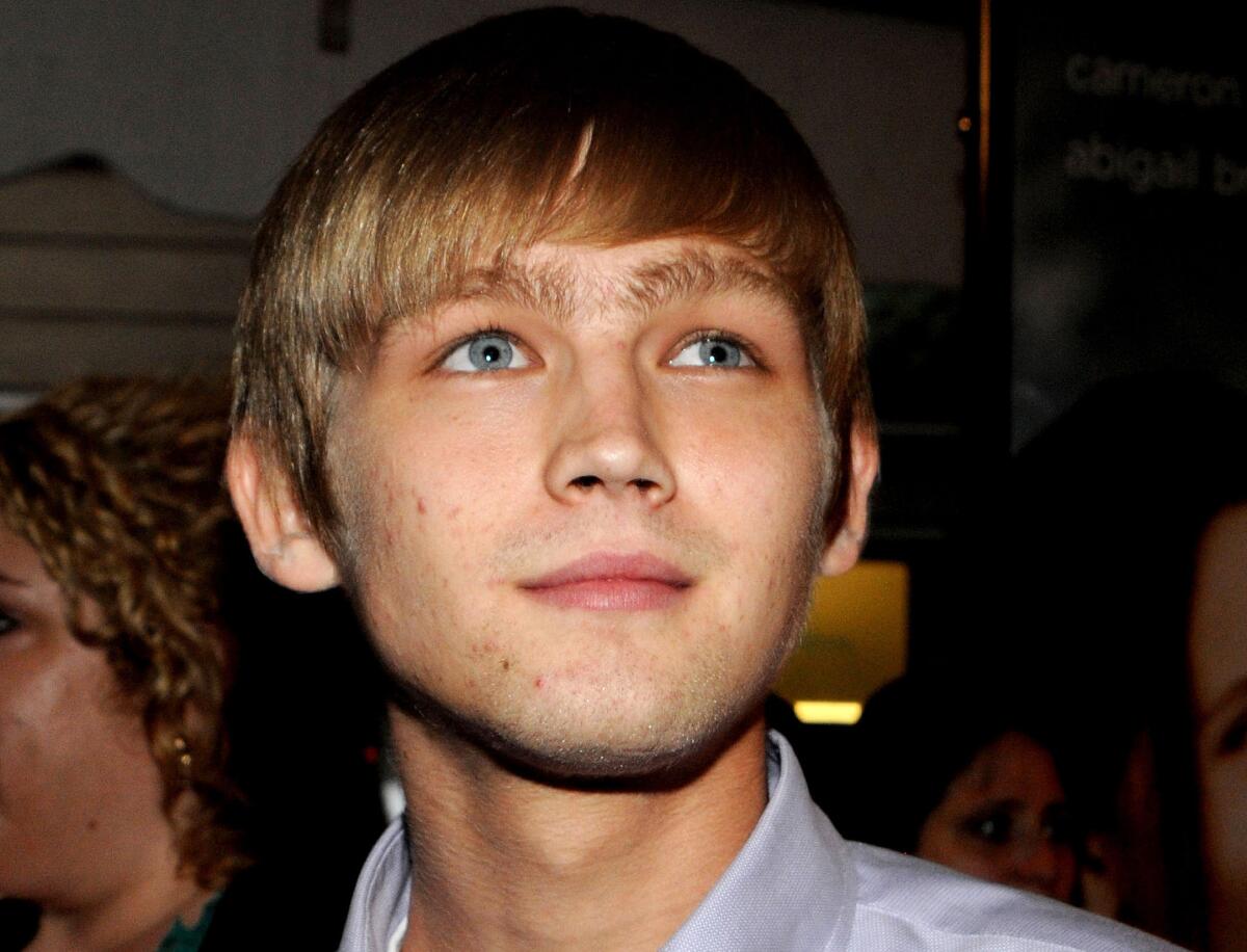 Closeup of a slightly smiling blond young man in a blue button-down shirt. 