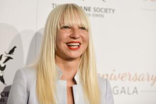 Sia is smiling while posing for photos while wearing an off-white blazer with long blonde hair down with bangs