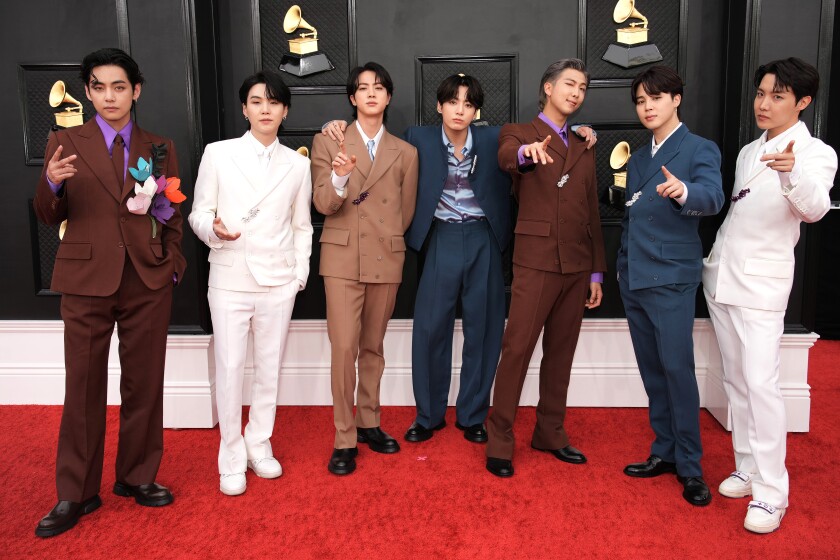 Seven men posing on a red carpet in alternating brown, white and blue suits