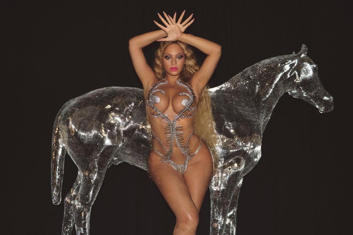 A woman with long blond hair posing in a silver monokini with her hands over her head in front of a translucent silver horse
