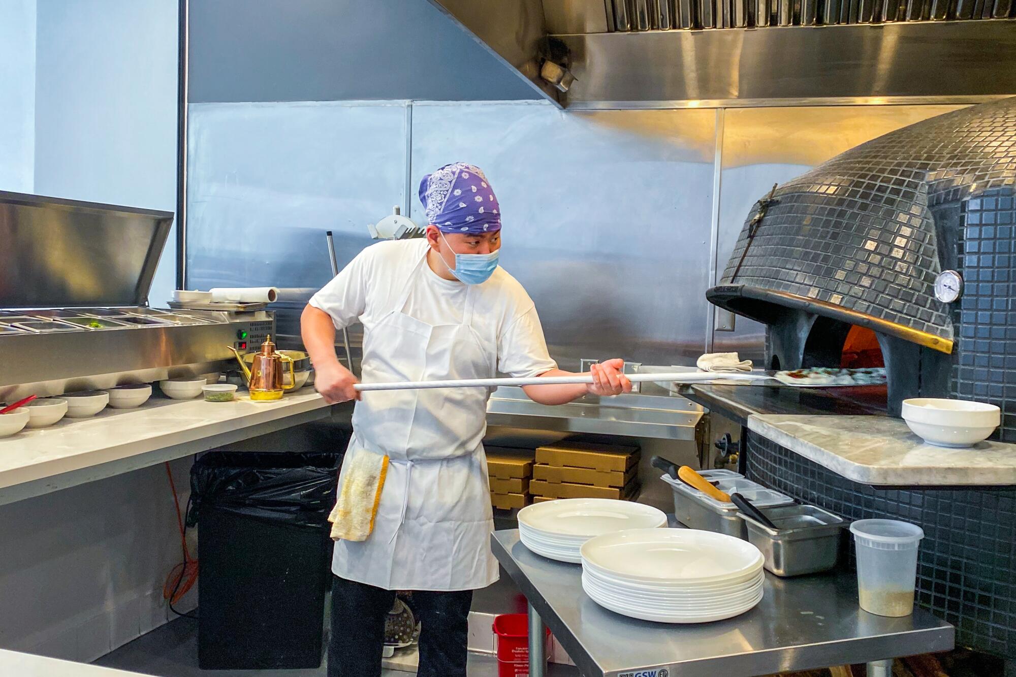 A chef in a face mask uses a peel to pull pizza from the oven at Pizzeria Sei.