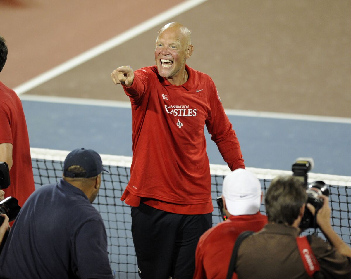 A man standing on a tennis court smiles and points to another person.