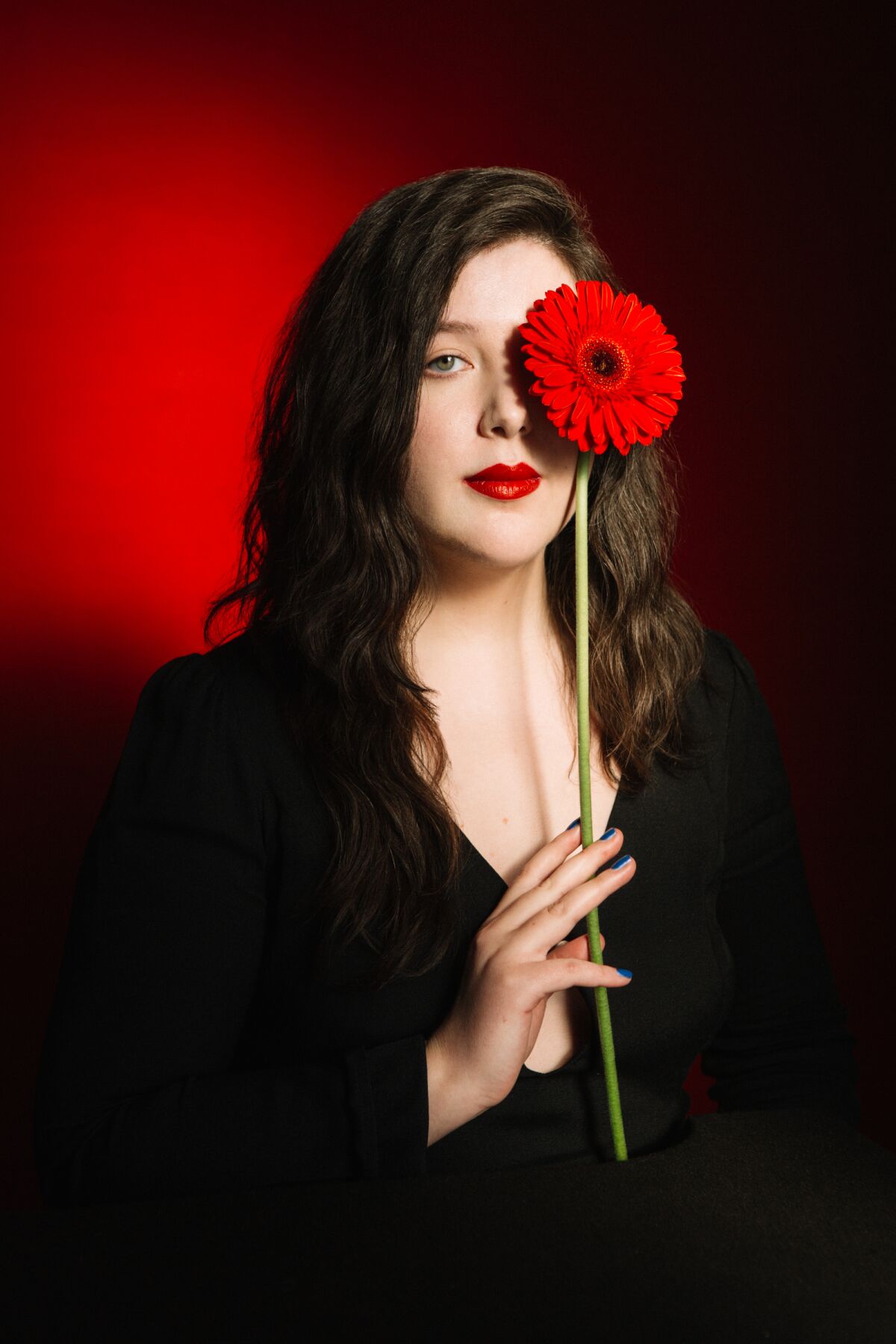 A portrait of Lucy Dacus holding a red flower in front of one eye.