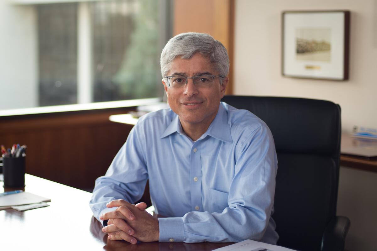 A man with gray hair, in glasses and a pale blue shirt, smiles while seated at a desk with hands clasped