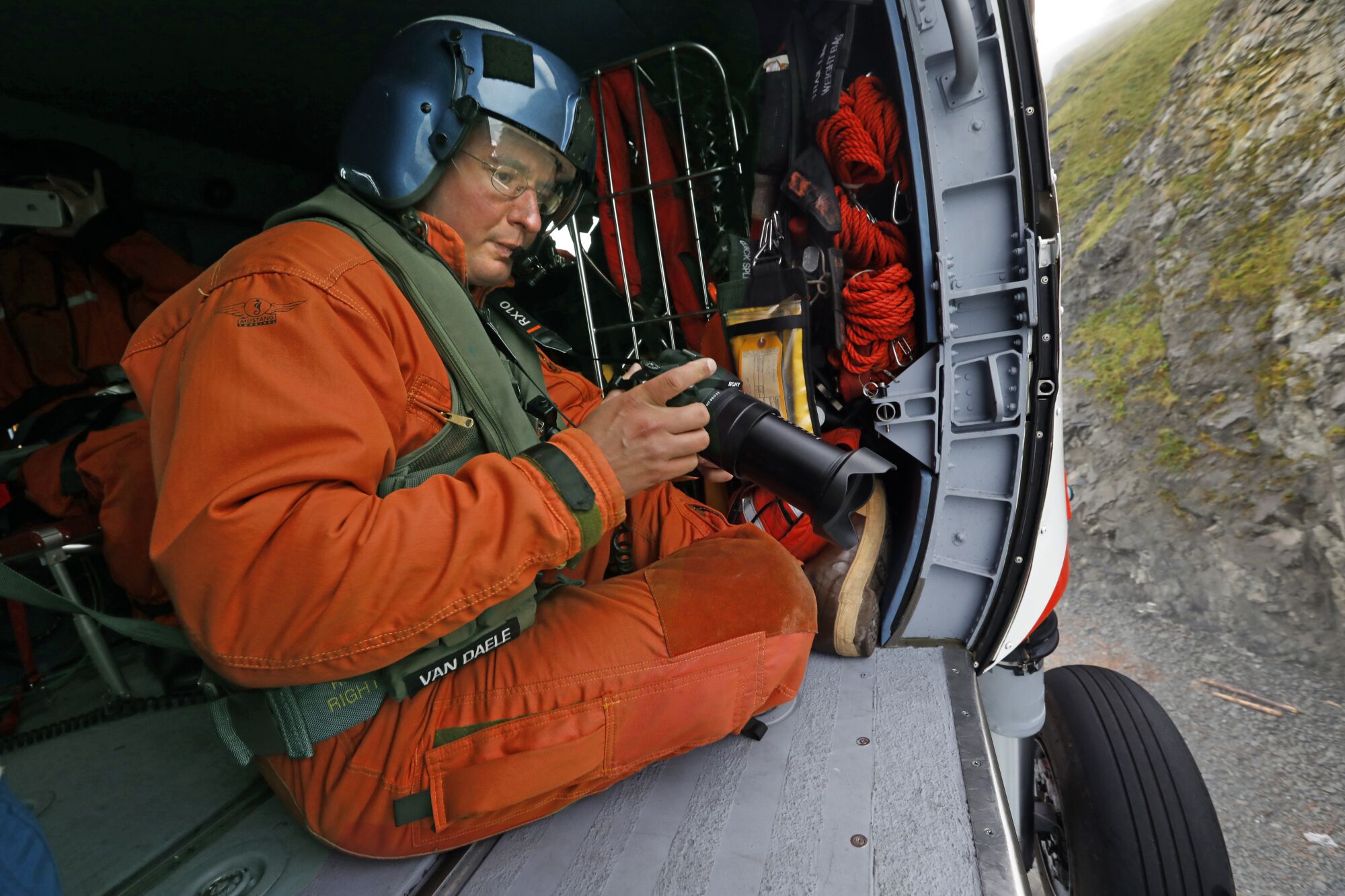 A man in an orange jumpsuit holding a camera as he scans the beaches below through the open door of a helicopter