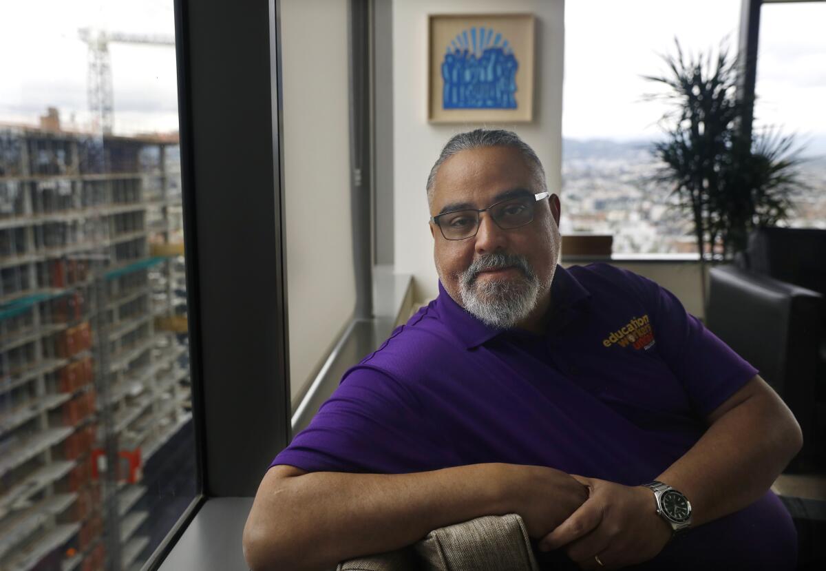 A seated man in a purple polo shirt poses near office windows with a city view