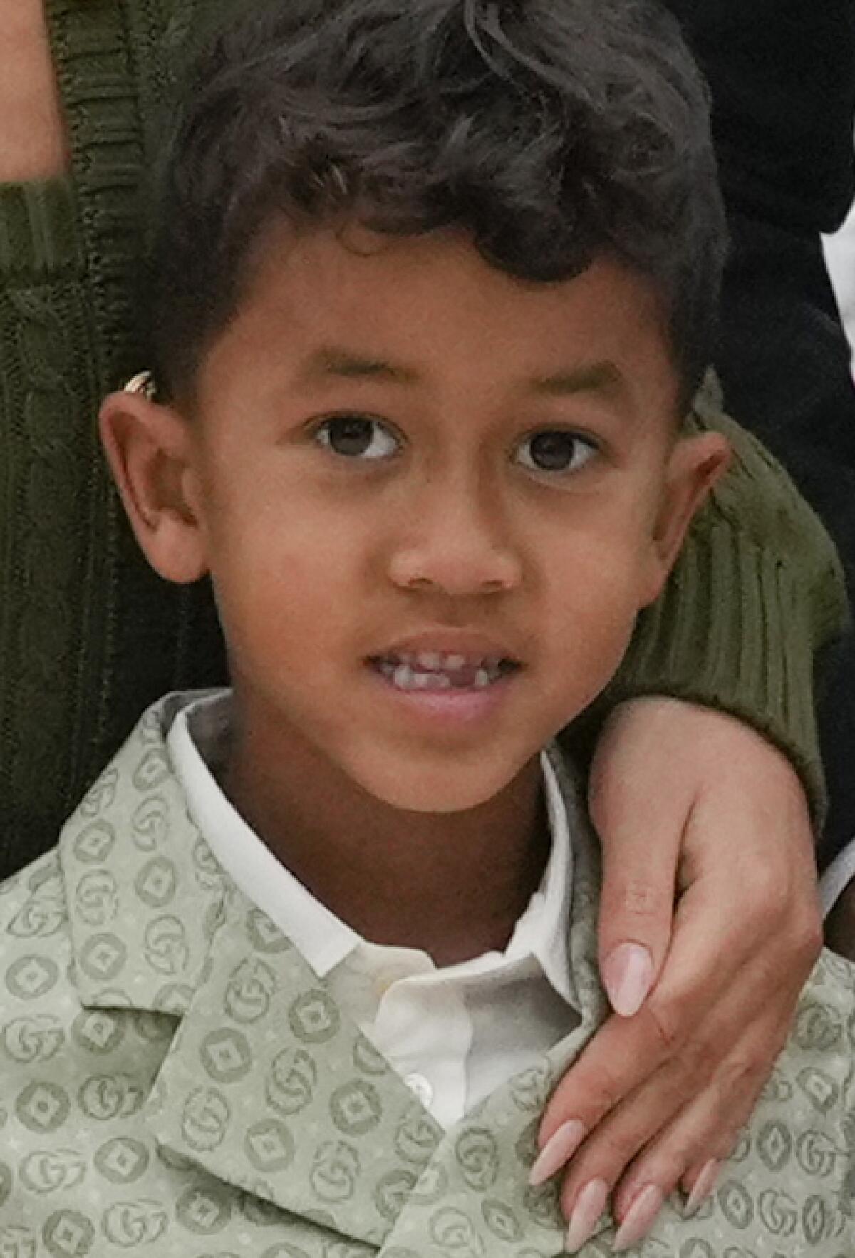 A boy with dark hair and missing teeth posing in a pale green jacket and white shirt with his mom's hand on his shoulder