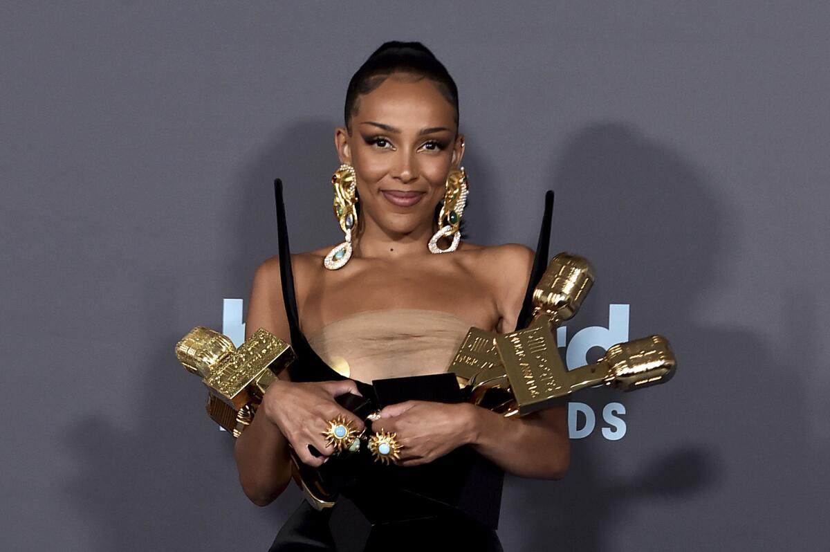 A woman wearing large gold earrings smiles while holding three trophies