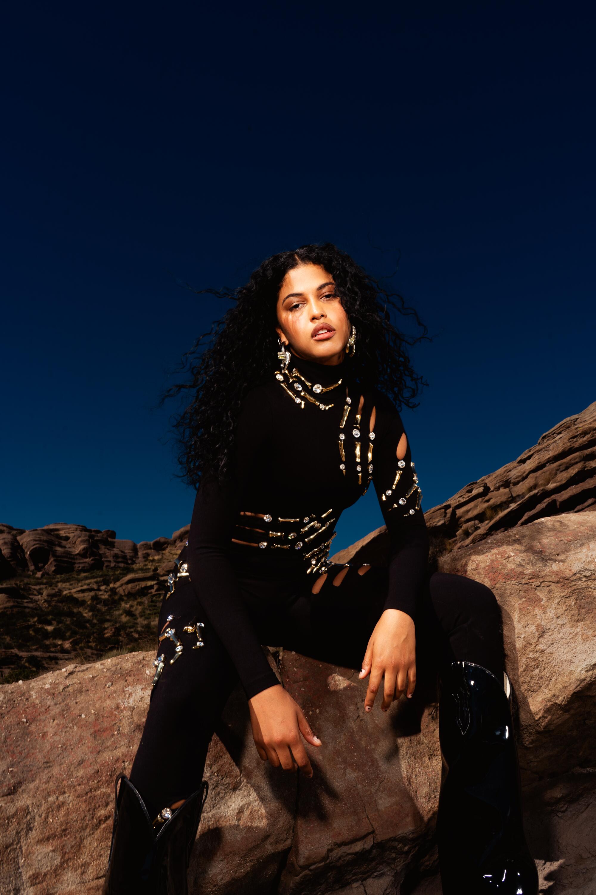 Artist Yendry sits on a rock in the desert in an all black outfit.