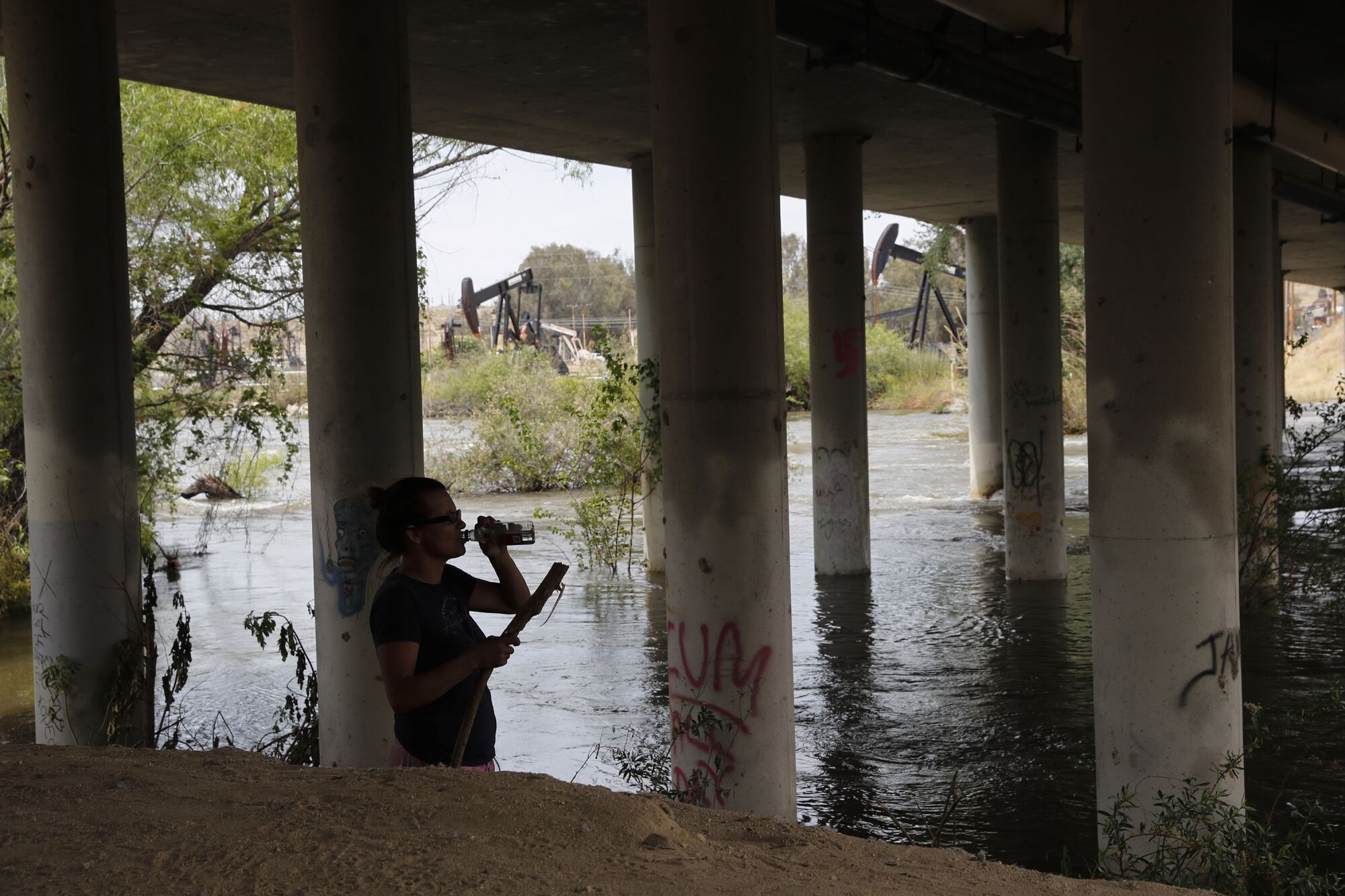 A woman drinks from bottle on a river bank beneath a bridge.