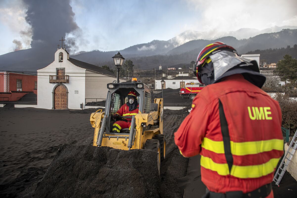 Military Emergency Unit personal clear black ash from volcano as it continues to erupt lava behind a church on the Canary island of La Palma, Spain on Wednesday Oct. 13, 2021. A new lava stream from an erupting volcano threatened to engulf another neighborhood on its way toward the Atlantic Ocean. Island authorities have ordered the evacuation of around 800 people from a section of the coastal town on Tuesday after the lava took a new course and put their homes in its probable path of destruction. (AP Photo/Saul Santos)